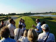 Thirteen participants standing on an embankment are listening to Manfred Meine at a field trip to Kreetsand