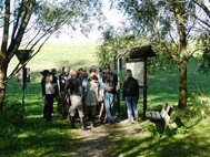 15 workshop-participants on a field trip in Heuckenlock are standing in front of a guide. The guide is using an information board to explain his topic to the audience
